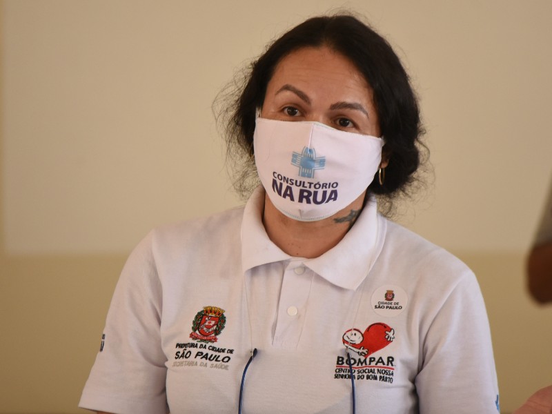 Samira tem cabelos e olhos castanhos. Ela usa uma máscara branca onde está escrito Consultório na Rua. Veste  camiseta branca com o emblema da Prefeitura de São Paulo do lado esquerdo e o logotipo do programa Consultório na Rua do lado direito